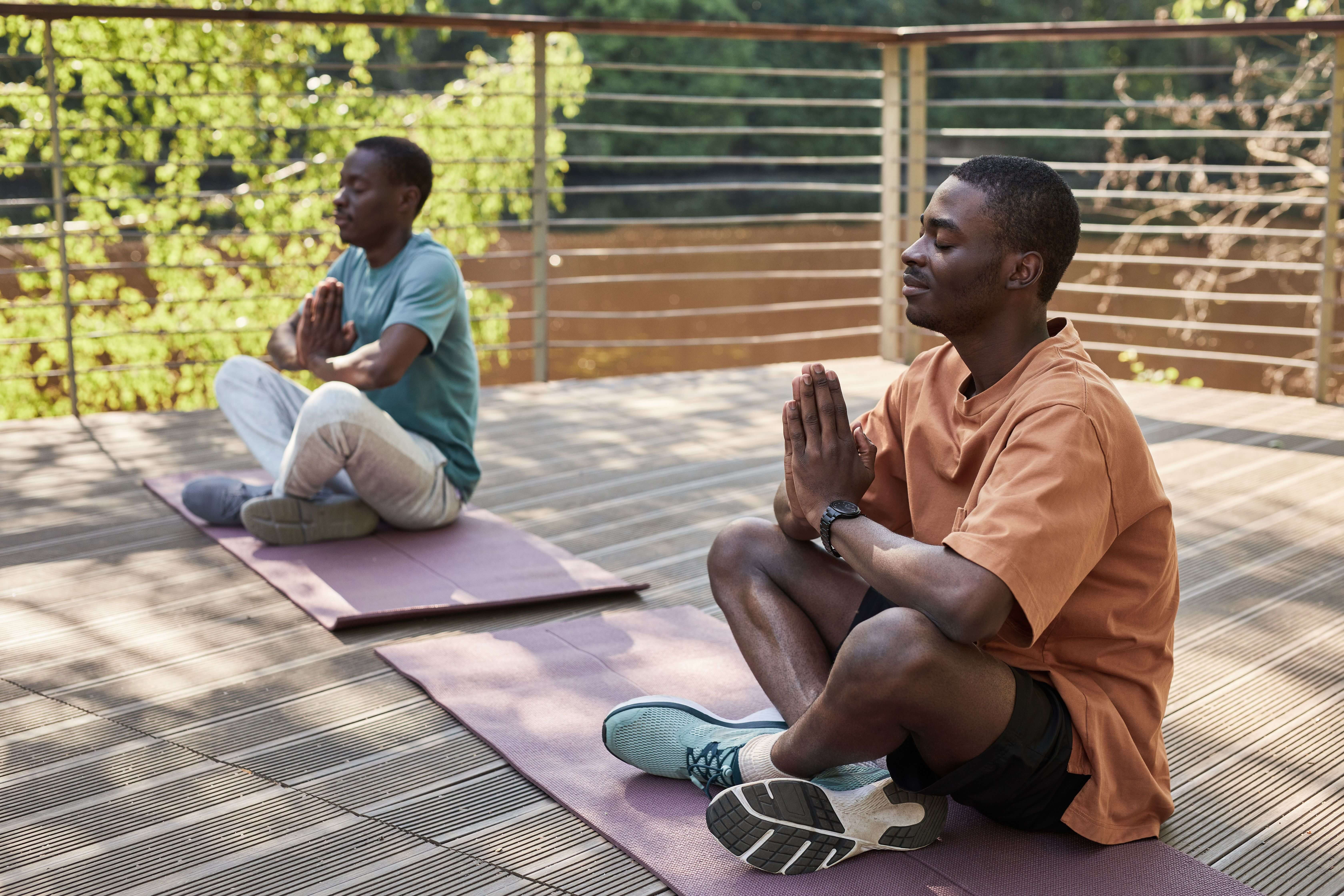 Two black men meditating outdoors on yoga mats in a serene setting.